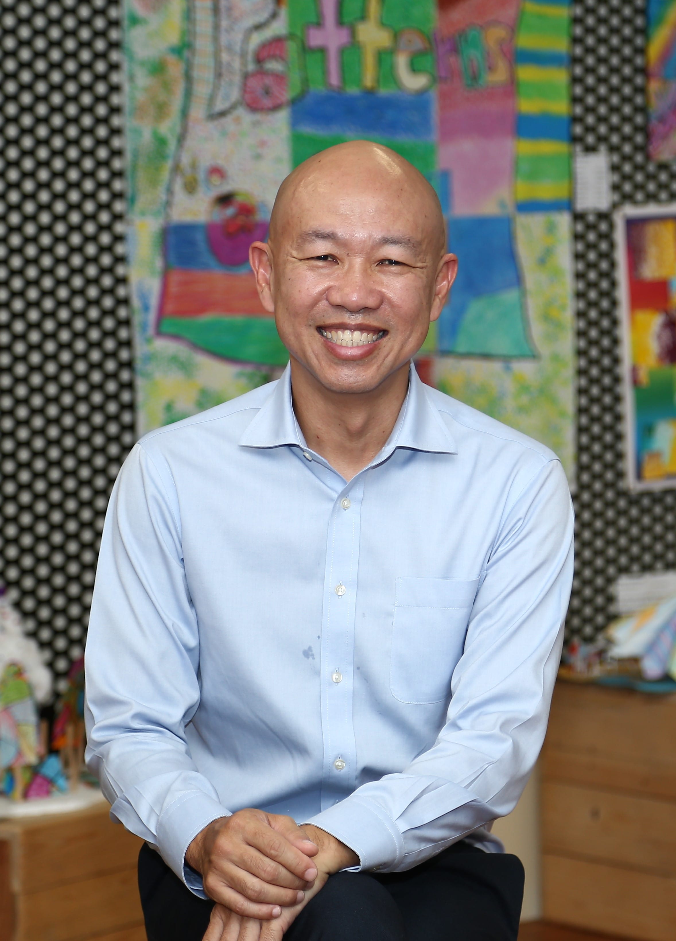 Smiling bald man in a light blue dress shirt seated with hands clasped before colorful children's artwork.